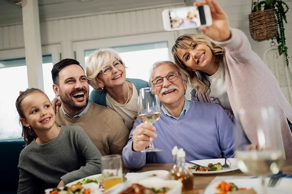 Família reunida à mesa a sorrir para uma fotografia, ilustrando a confiança e a qualidade de vida que uma prótese fixa sobre implantes pode proporcionar no dia a dia.